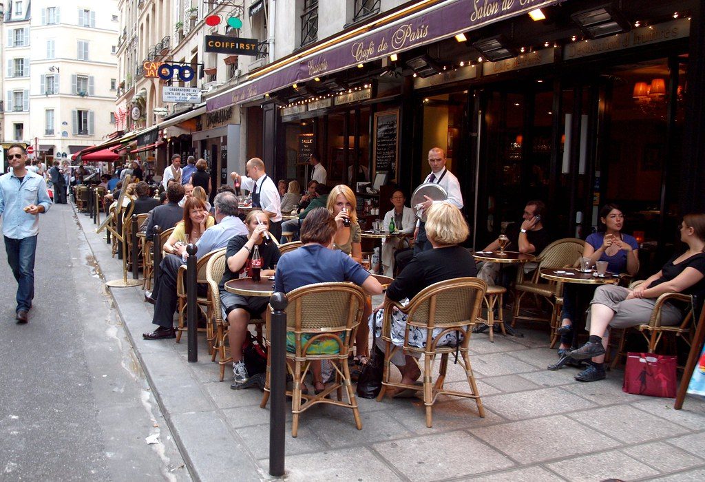 People sitting at small round tables outside a Paris cafe on a sunny street.